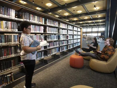 A child is standing in the aisle of the bookshelves talking to 2 other children sitting in chairs and holding books