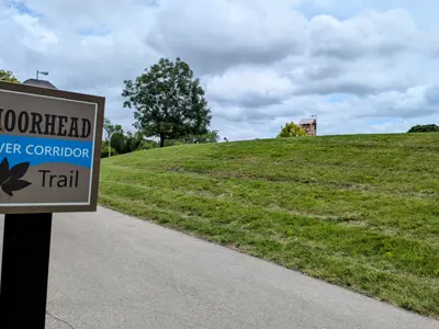 A sign that says Moorhead River Corridor Trail with a park trail behind it